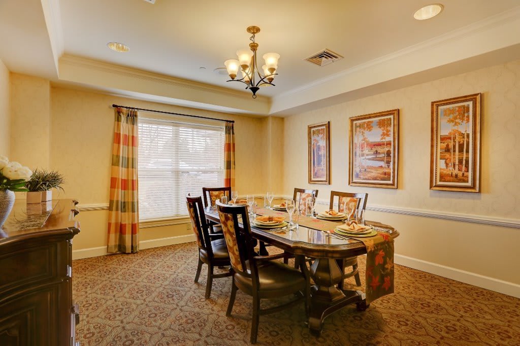 A cozy dining room with a chandelier, wooden table and chairs, framed art on the walls, and a large window with curtains.