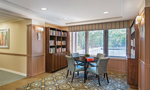 A cozy reading area featuring a wooden bookshelf filled with books, a round table surrounded by chairs, and plenty of natural light from large windows.