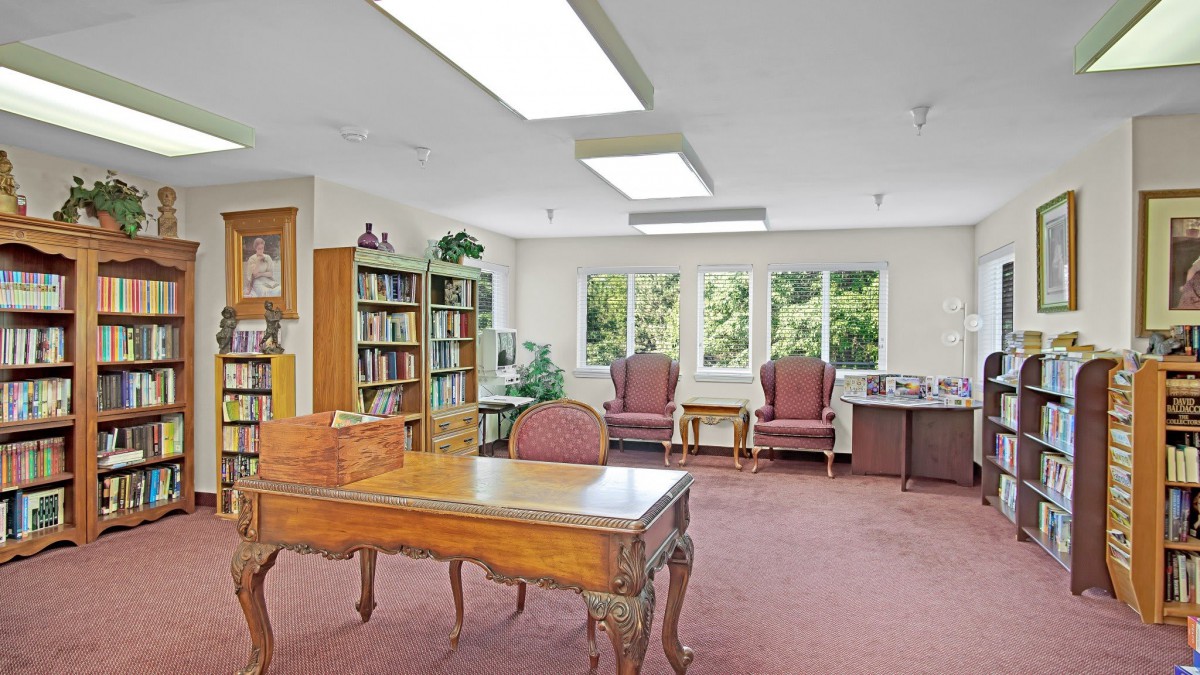 A cozy office space with a wooden desk, chairs, and bookshelves filled with books, complemented by large windows and indoor plants.