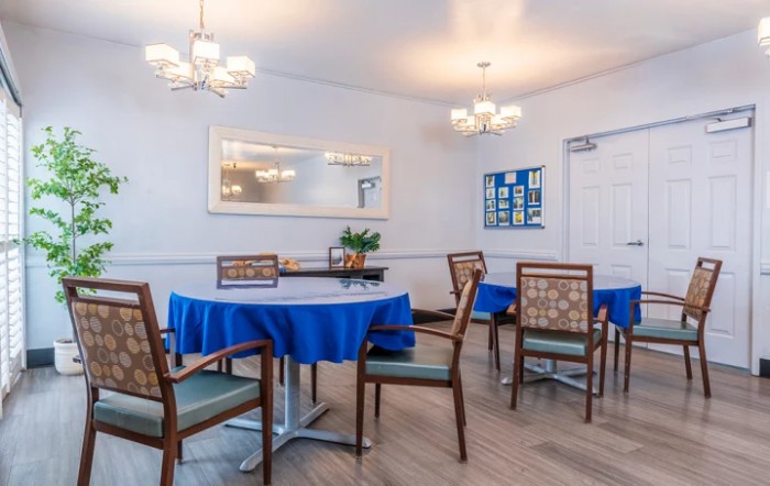 A cozy dining area with tables covered in blue tablecloths and surrounded by wooden chairs, illuminated by elegant chandeliers.