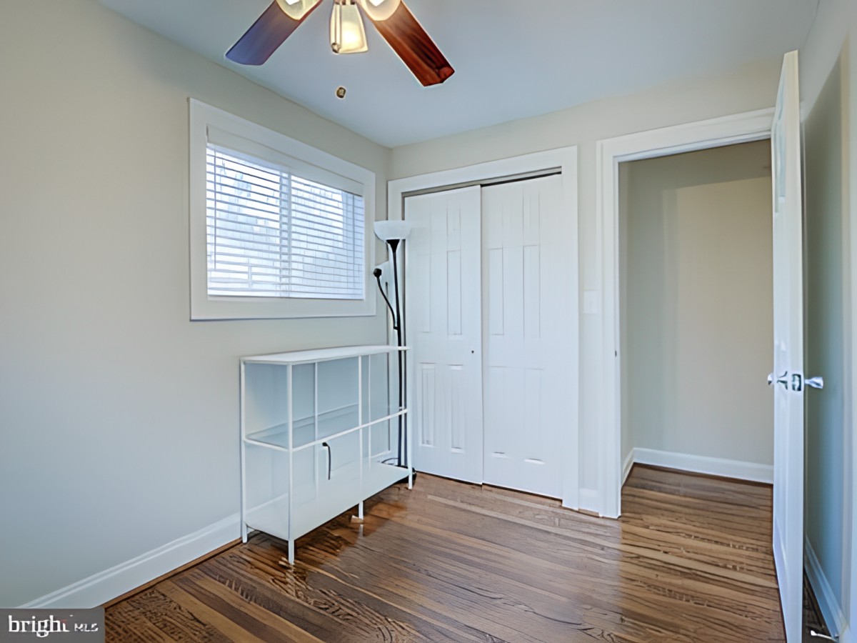 A simple bedroom interior with wooden flooring, a closet with sliding doors, and a ceiling fan.