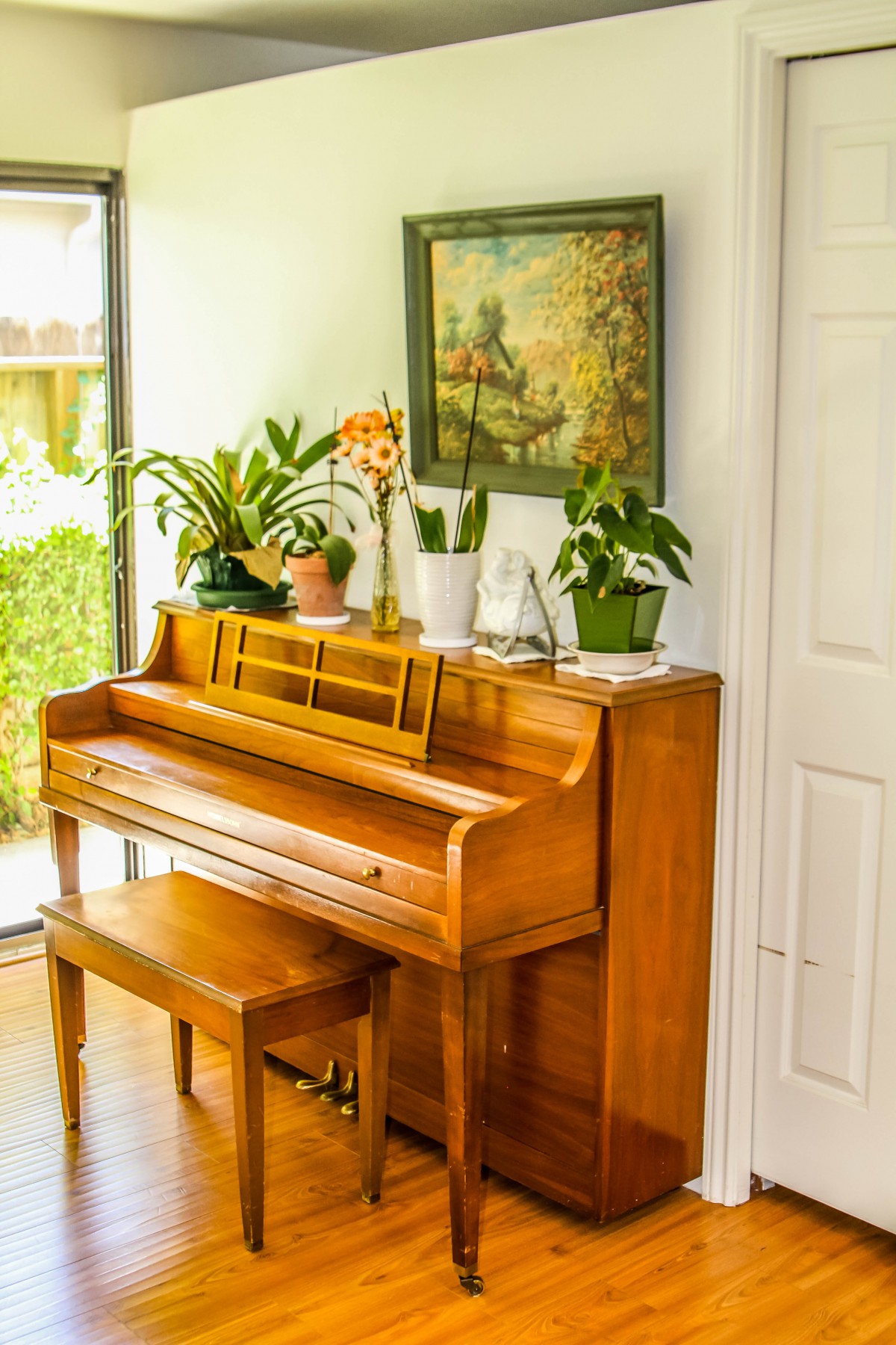 A cozy interior featuring a wooden piano adorned with potted plants and artwork, illuminated by natural light from a nearby window.