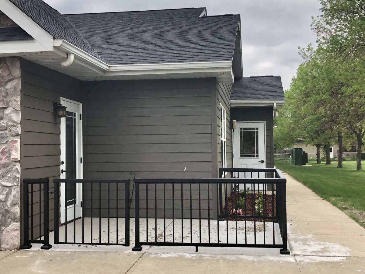 A modern house exterior with a small patio area enclosed by black railings and adjacent sidewalk.