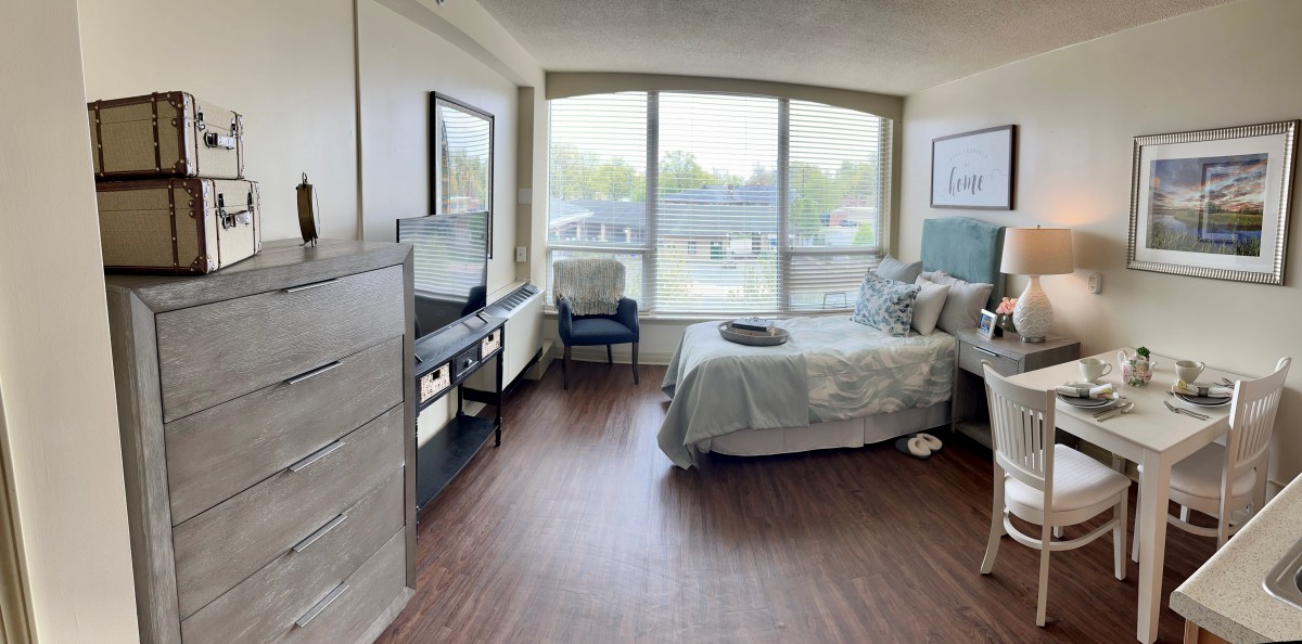 A modern bedroom with large windows allowing natural light, featuring a bed, dresser, and TV setup.