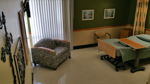 A view of a cozy room featuring an armchair and a hospital bed, with natural light streaming in through the window.