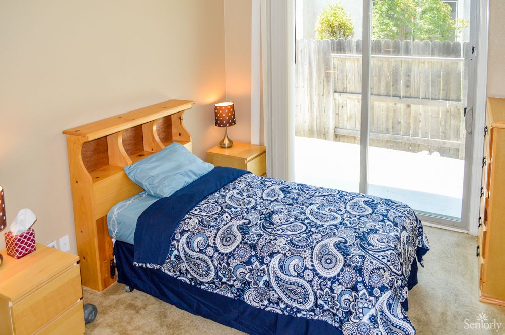 A cozy bedroom with a single bed featuring blue paisley-patterned bedding next to a nightstand and lamp, illuminated by natural light through a sliding door.