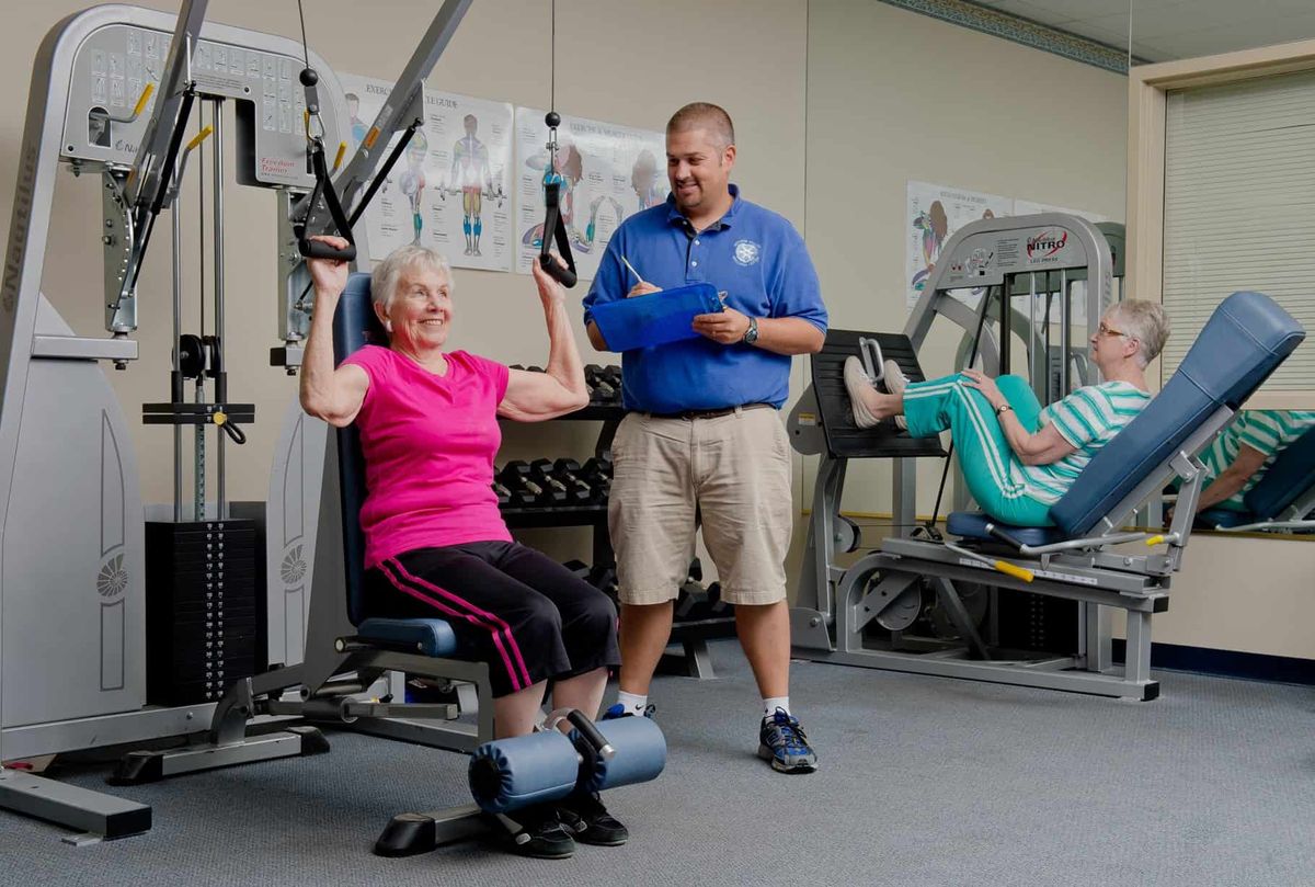 An elderly woman is working out on a gym machine with guidance from a trainer.