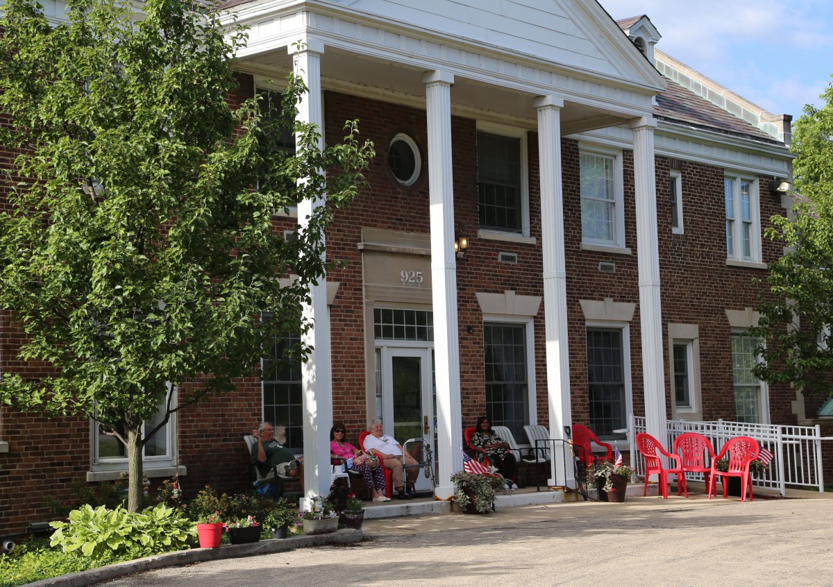 A brick building with large white columns and a porch decorated with chairs and plants.