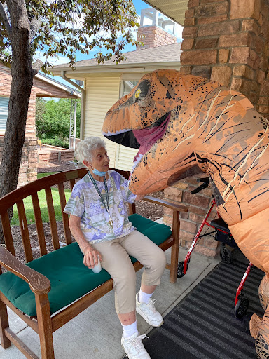 An elderly person shares a playful moment with someone in a dinosaur costume on a sunny day.