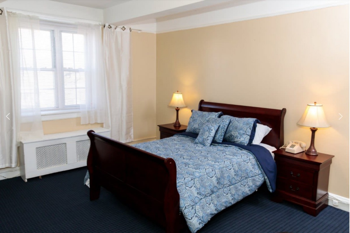 A cozy bedroom with a wooden bed, a bedside lamp, and a window with white curtains.