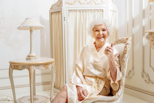 An elderly woman in a robe, smiling while sitting in a luxurious room.