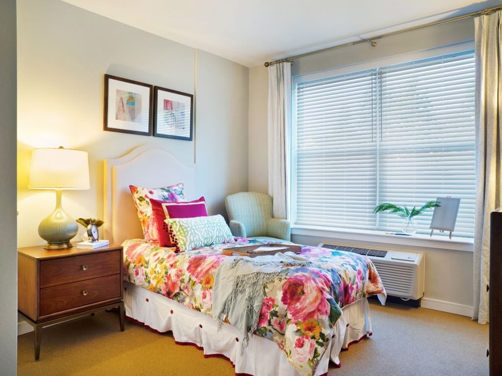 A cozy bedroom with floral bedding, a side table with a lamp, and a large window with blinds.