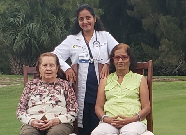 A doctor stands behind two seated elderly women outdoors.