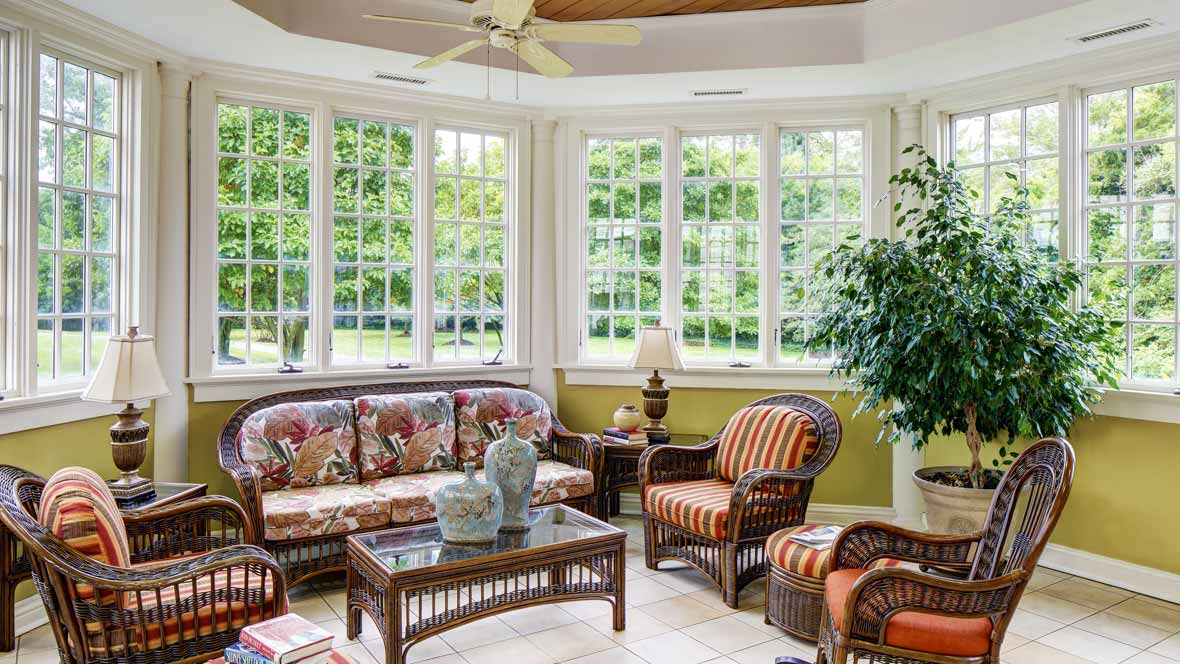 A bright sunroom with wicker furniture, large windows, and lush greenery.