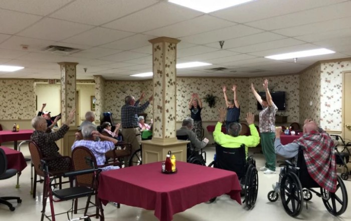 A group of elderly individuals participating in a seated exercise session, some using wheelchairs, in a communal setting.