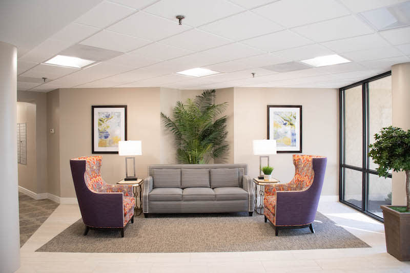 A cozy lobby area featuring a grey sofa and patterned chairs with lamps on side tables, framed artwork on the walls, and a large indoor plant.