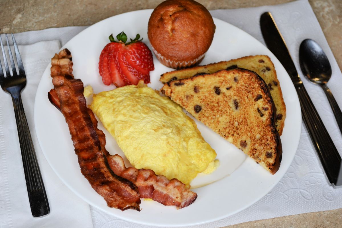 A hearty breakfast plate with an omelette, crispy bacon, toast, a muffin, and a fresh strawberry.