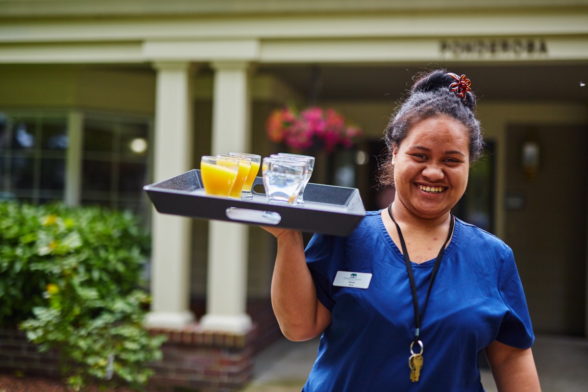A woman in a uniform is smiling and carrying a tray with drinks, including orange juice and water, outdoors.