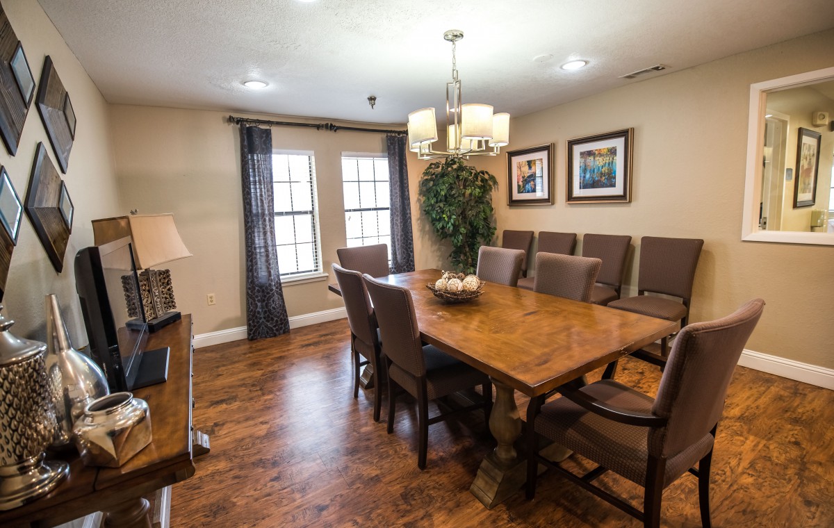 A cozy dining room with a large wooden table, surrounded by chairs and illuminated by a chandelier. The room features artwork on the walls, hardwood flooring, and natural light pouring in through double windows adorned with curtains.