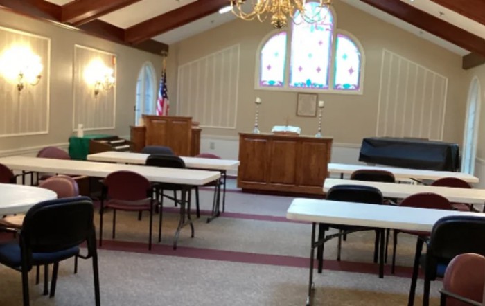Interior of a church meeting room with tables and chairs arranged, featuring a pulpit and stained glass windows.