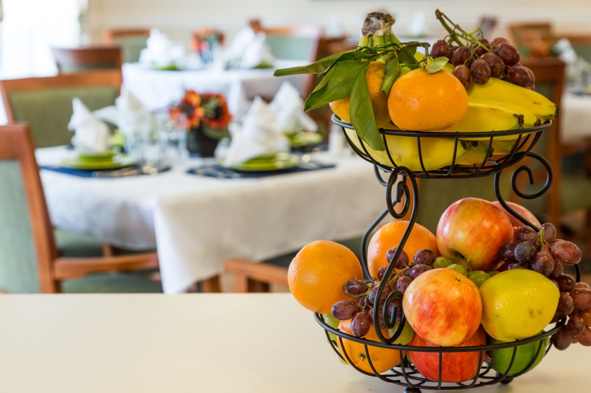 A fruit basket with oranges, peaches, and grapes sits in the foreground, while a neatly set dining table is blurred in the background.