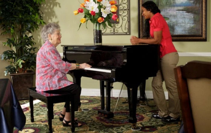 An elderly woman plays the piano while a caregiver listens attentively.