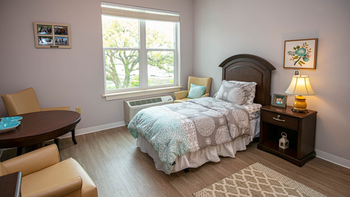 A cozy bedroom with a single bed, wooden floor, and ample natural light coming through a large window.