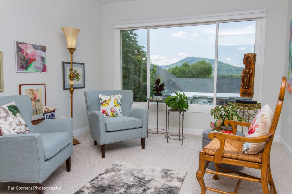 A cozy living room with blue armchairs, a large window offering a scenic view, and various plants adding a touch of greenery.