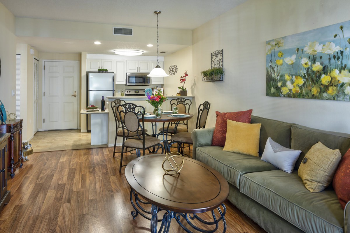A cozy living and dining area with a view into the kitchen, featuring a sofa, an oval coffee table, and a dining set with decorative cushions.