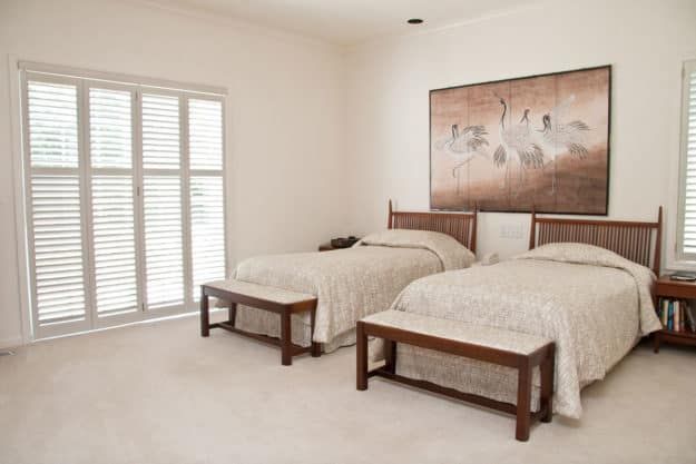 A neatly arranged bedroom featuring twin beds with a central painting and window blinds.