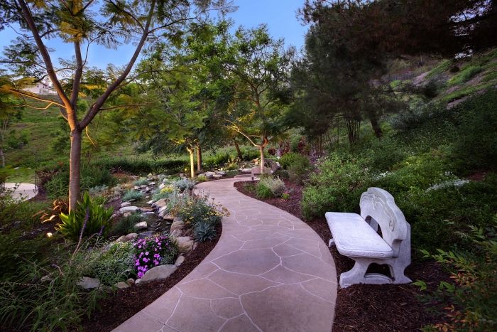 A peaceful garden pathway with a stone bench surrounded by trees and colorful flowers.