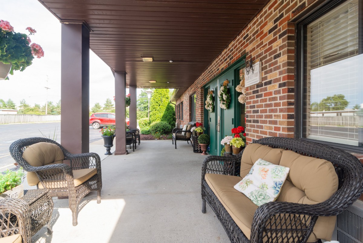 A cozy porch with wicker furniture and decorative plants outside a brick building.