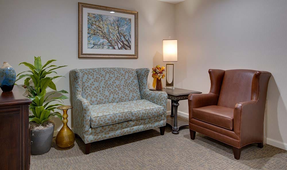 A cozy living room with a patterned sofa, brown armchair, side table, and a lamp under a framed painting.