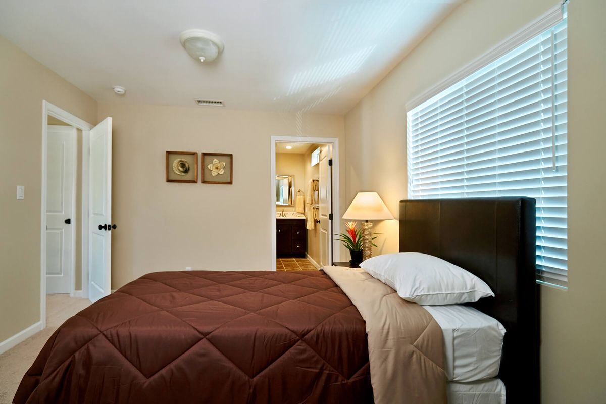 A cozy bedroom with a neatly made bed, bedside lamp, and a window letting in natural light.