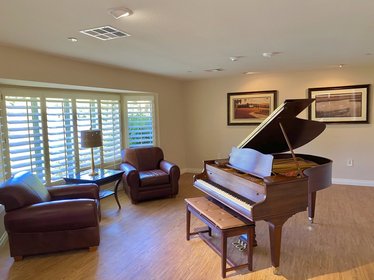 A cozy living room featuring a grand piano, comfortable armchairs, and warm natural light filtering through wooden shutters.
