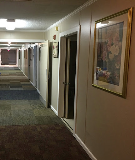 A dimly lit hotel hallway featuring carpeted floors, doors leading to rooms, and a framed artwork on the wall.