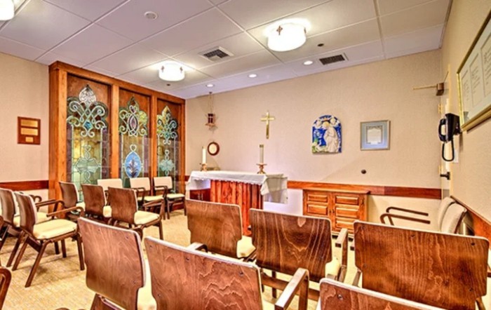 A serene chapel interior with rows of chairs facing an altar, adorned with decorative windows and soft lighting.