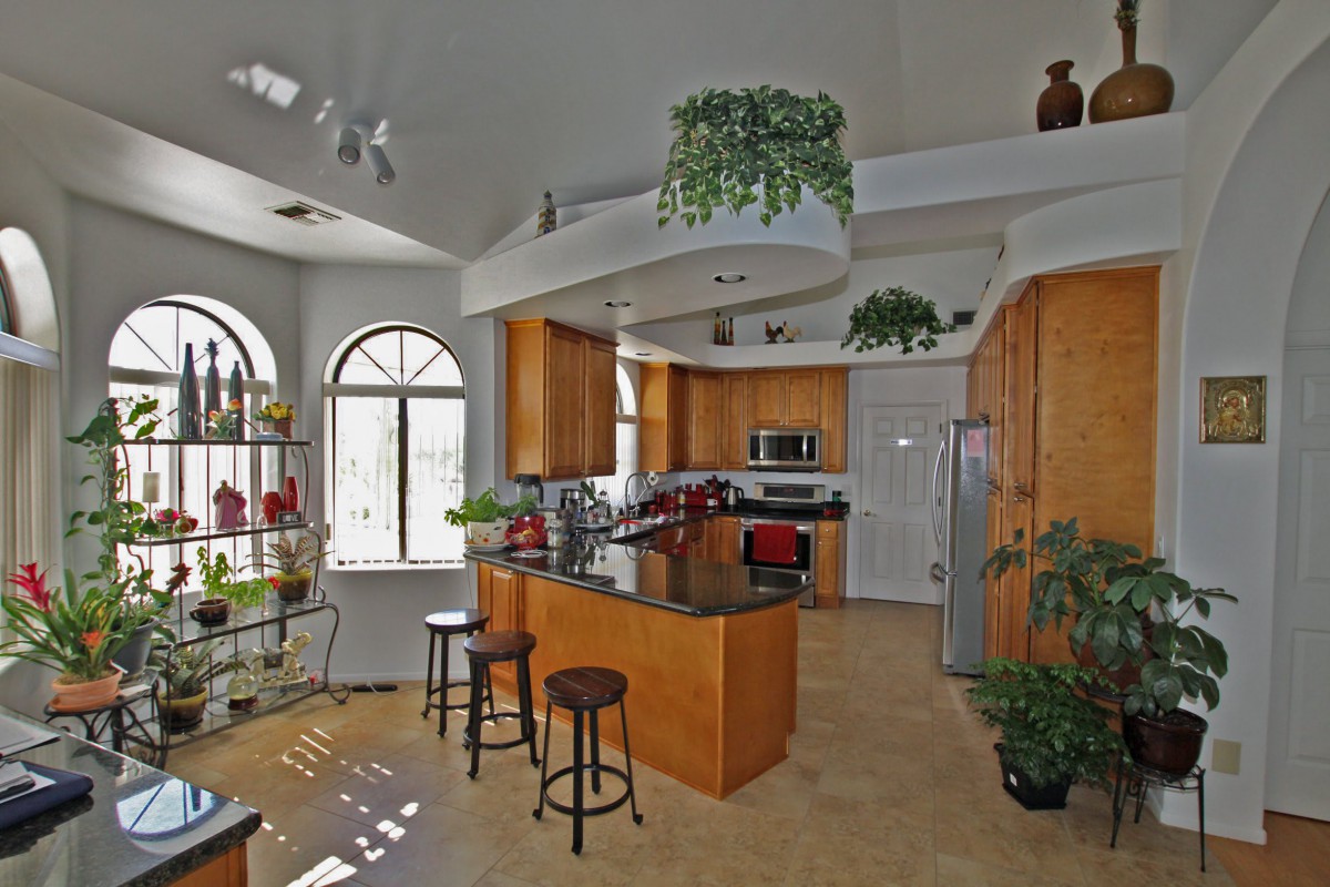 A modern kitchen featuring wooden cabinets, a central island with bar stools, and stainless steel appliances, enhanced by natural light and greenery.