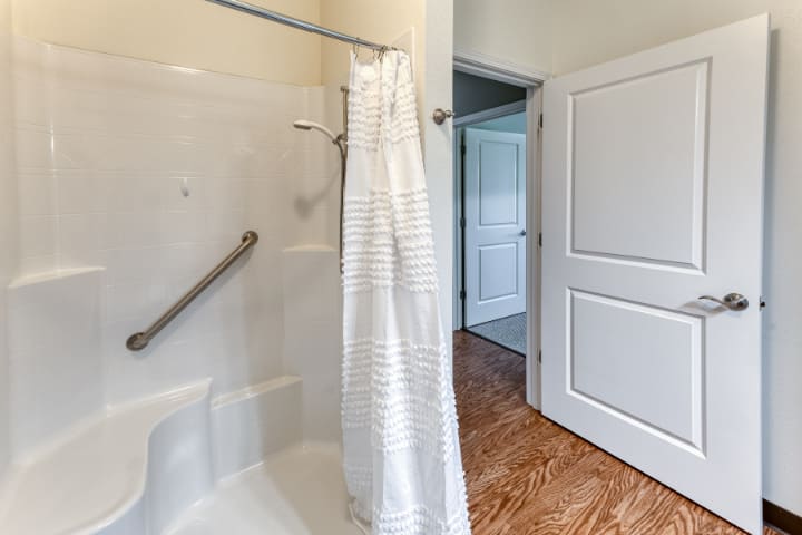A bathroom featuring a shower with a curtain and handrail, opening into a room with hardwood floors.