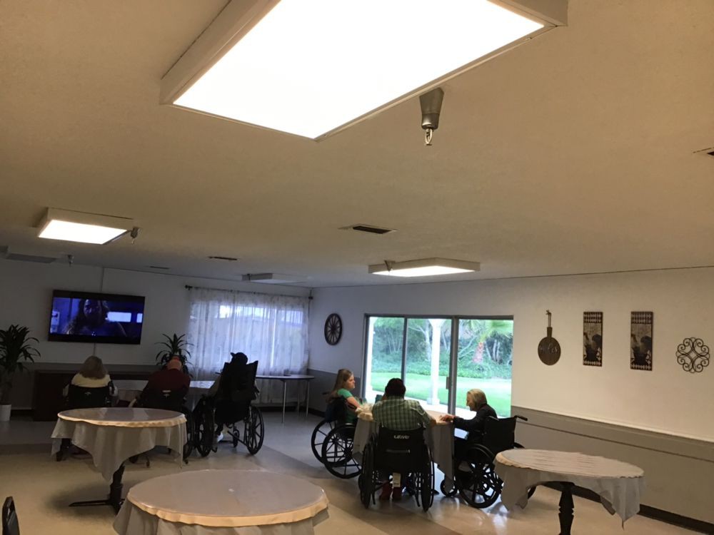 A nursing home common area with seniors in wheelchairs watching television.