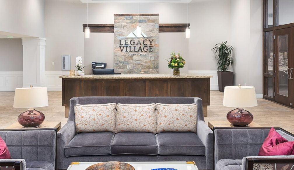 A modern lobby area featuring a comfortable couch, decorative plants, and a reception desk with the sign 'Legacy Village'.