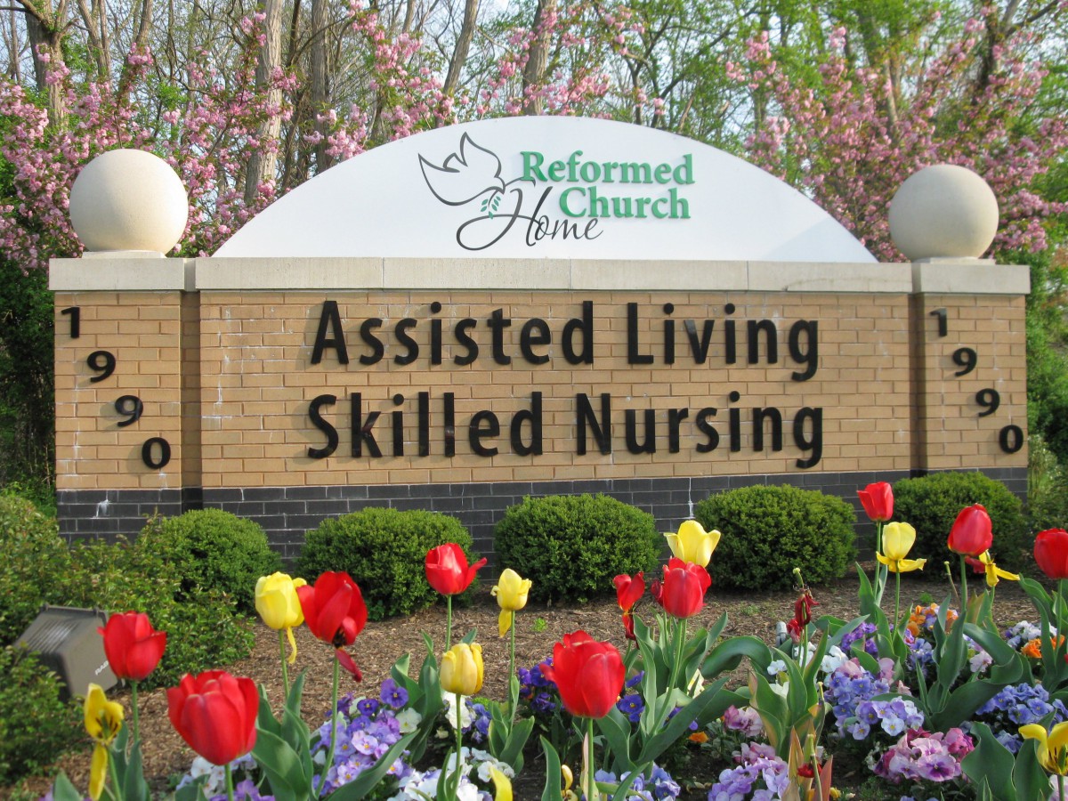 Sign for a Reformed Church Home, offering Assisted Living and Skilled Nursing services, surrounded by colorful spring flowers.