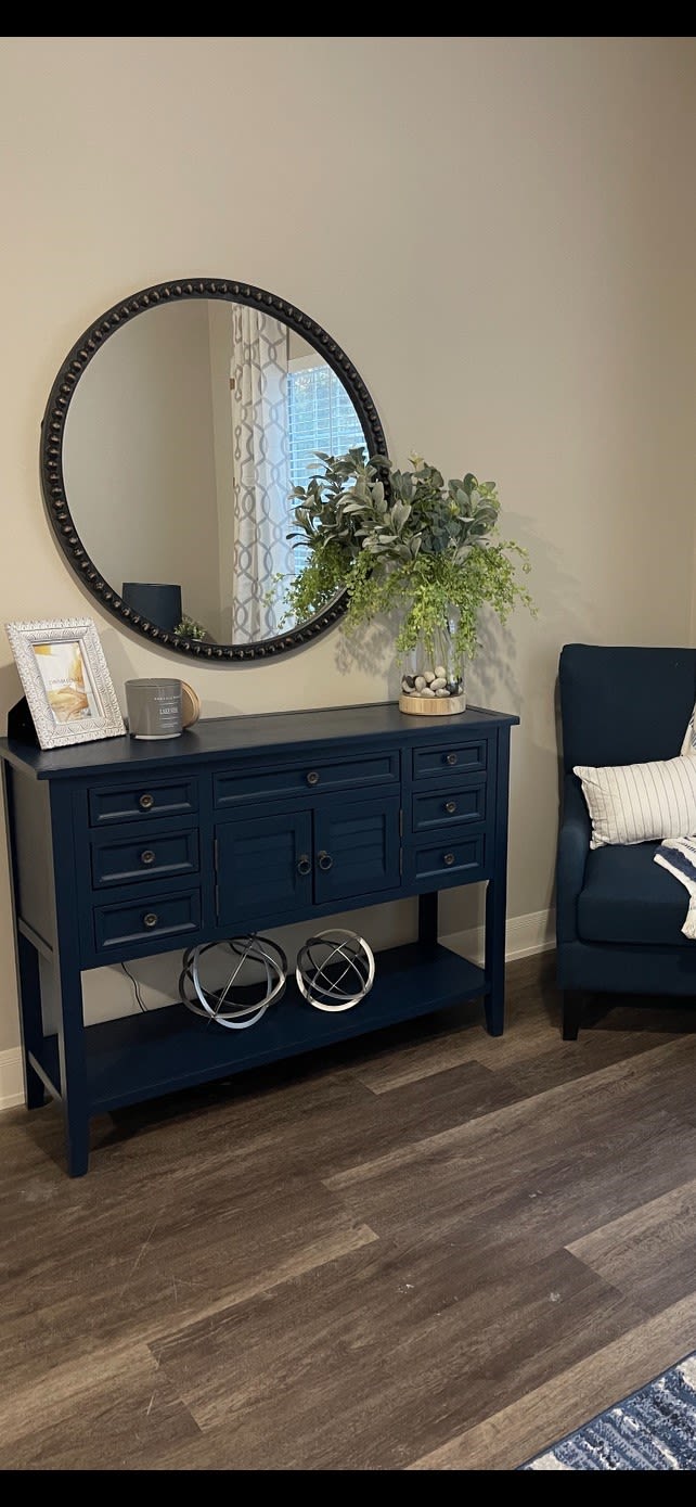 A stylish living room corner featuring a navy blue sideboard with decorative items, a round mirror, and a cozy armchair.