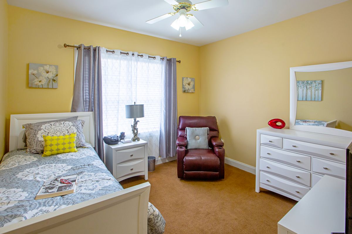 A cozy bedroom with yellow walls, featuring a bed, armchair, dresser, and a window with curtains.