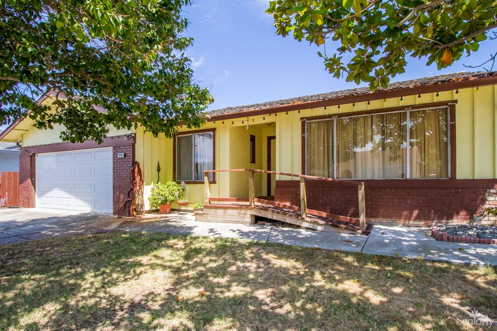 A yellow house with a ramp and a tree in the front yard under a clear blue sky.