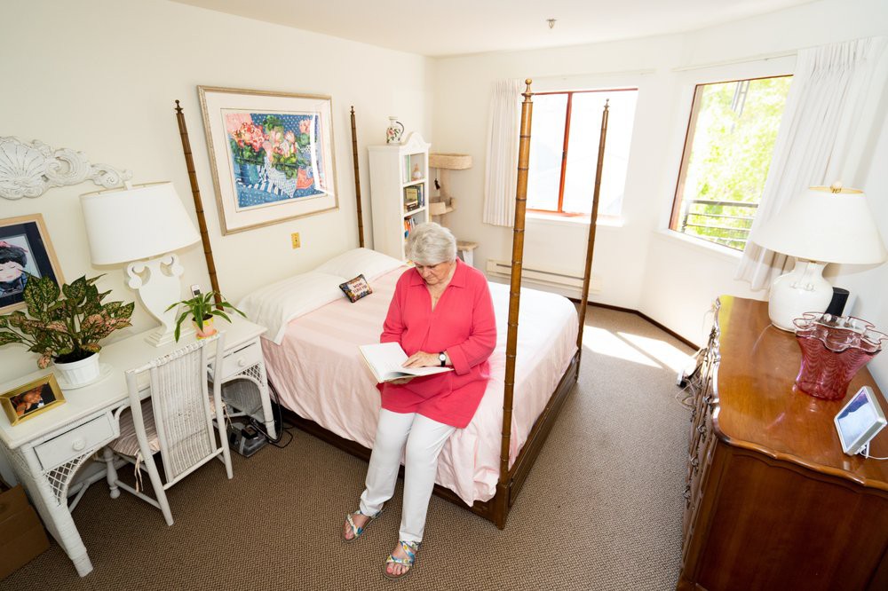 A woman sits on a bed in a brightly lit bedroom reading a book.