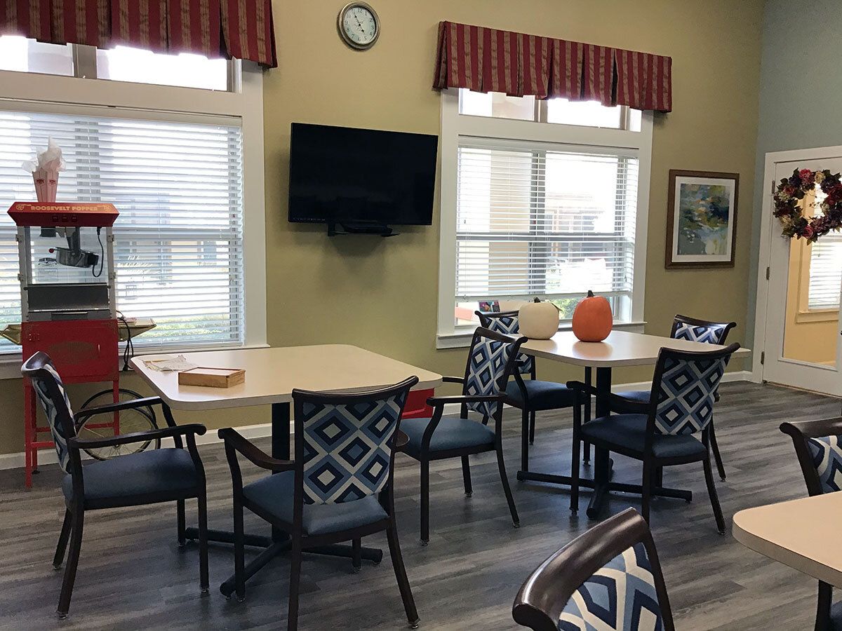 A cozy room with tables and chairs, featuring a wall-mounted television and large windows.