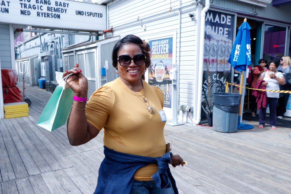 A woman wearing sunglasses and casual clothing smiles while walking on a boardwalk.