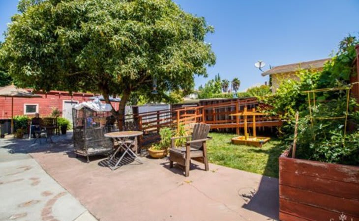 A sunny backyard featuring a patio area with outdoor furniture, surrounded by grass and a wooden fence.