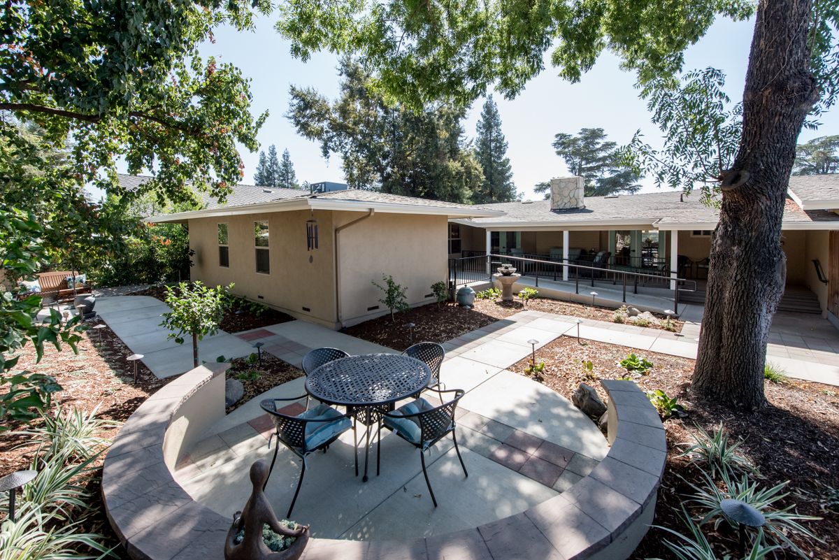 A modern patio area with a table and chairs surrounded by a landscaped garden and trees, adjacent to a house.